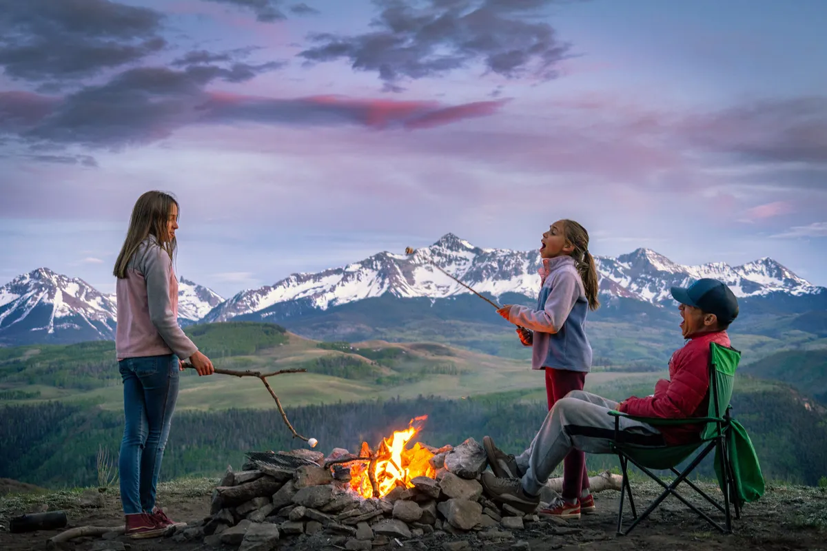 A lively, heartwarming outdoor scene of children roasting marshmallows over a campfire, with an adult seated nearby and awe-inspiring mountain scenery in the background. The group is immersed in nature, sharing stories, and enjoying the peaceful, joyful atmosphere of a family adventure.