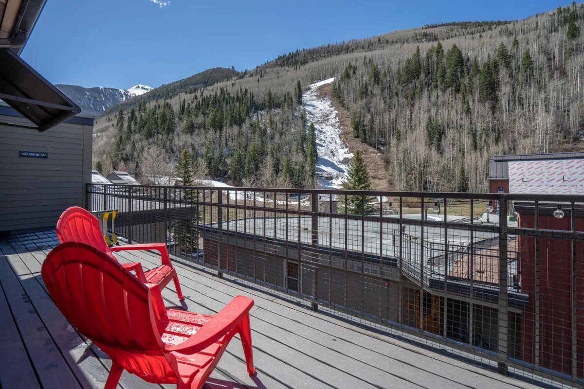 A tranquil exterior scene featuring red Adirondack chairs positioned to take in expansive mountain views with the ski slope visible beyond. The early morning or late afternoon light bathes the area, inviting relaxation and reflection after a day outdoors.