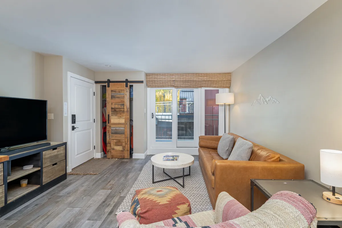 The image showcases a cozy living room with a plush leather sofa positioned beside a rustic wooden sliding door, a modern television mounted opposite, and a stylish coffee table in the center. The space invites guests to unwind, socialize, or enjoy a quiet evening indoors.