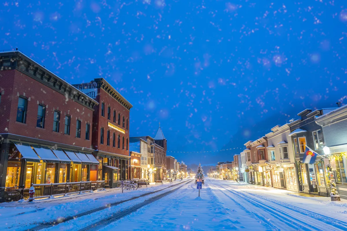 A snow-covered street lined with charming, decorated storefronts and a glowing Christmas tree, illuminated by streetlights. The scene evokes the warmth and excitement of Telluride’s winter festivities and community events.