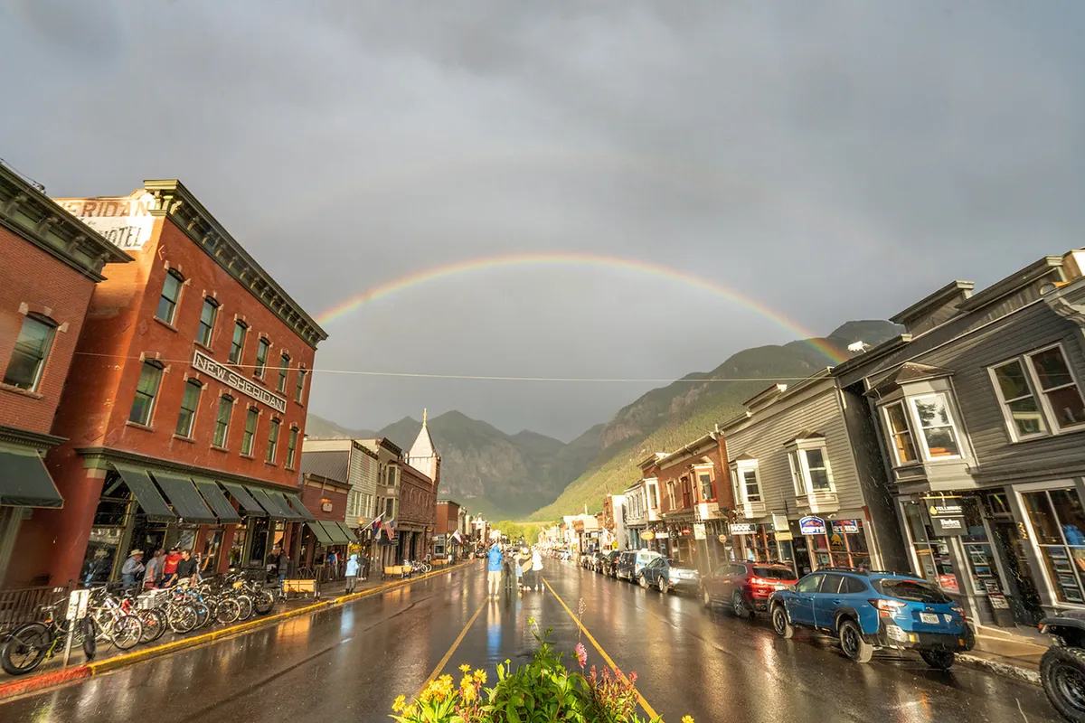A lively Telluride street scene with historic buildings, a vibrant rainbow arching overhead, wet street reflections, and bicycles parked along the curb. The area is bustling and cheerful, capturing the spirit of town life in the summer.