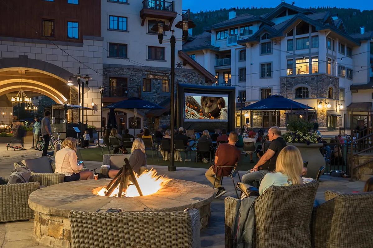An outdoor fire pit glows as guests gather around, enjoying an outdoor movie under the stars. Comfortable lounge chairs and a warm fire create a cozy, communal atmosphere.