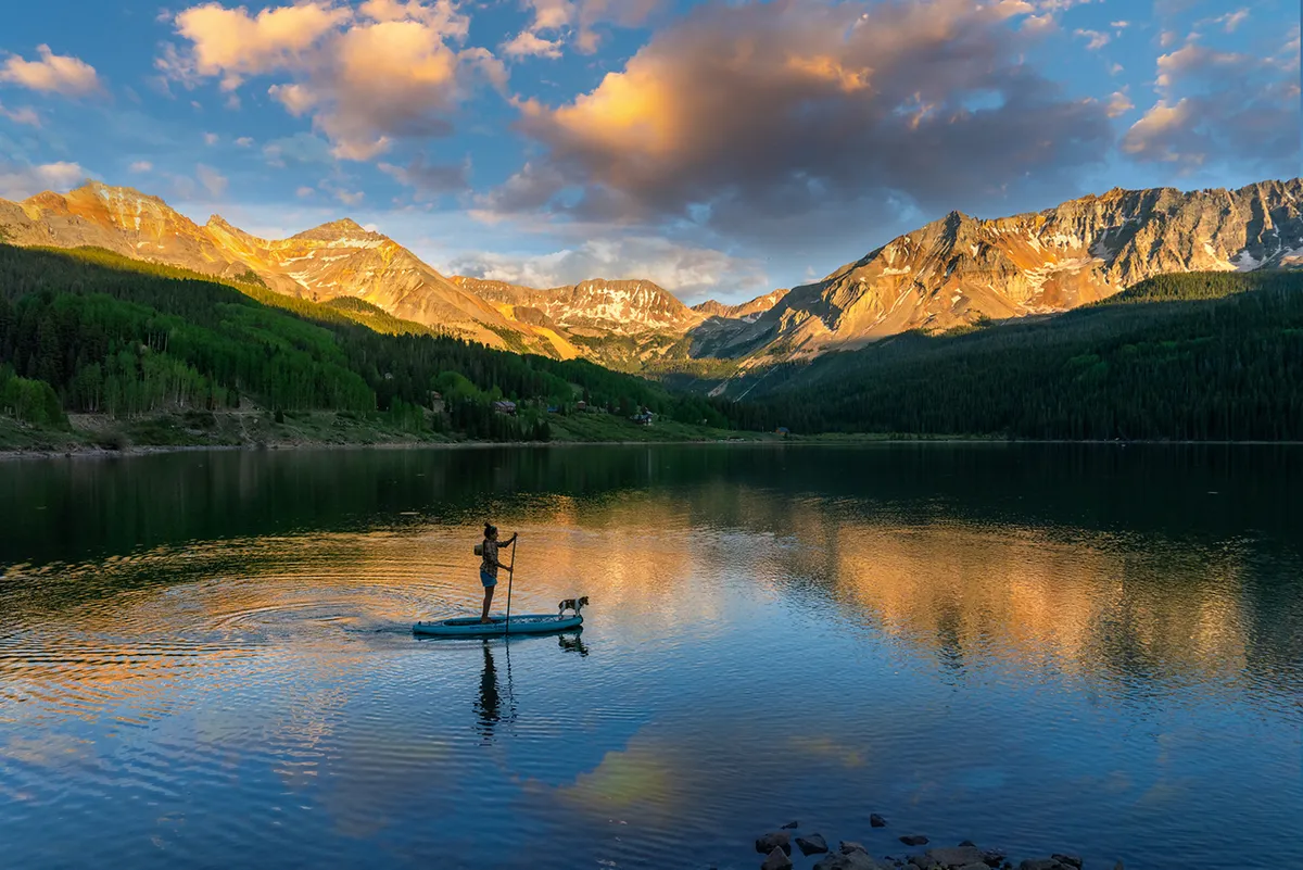 A serene lake scene with a person paddle-boarding alongside a dog, backed by majestic mountains and mirrored by still water. The image evokes the tranquility and sense of connection to nature that defines Telluride in the warmer months.