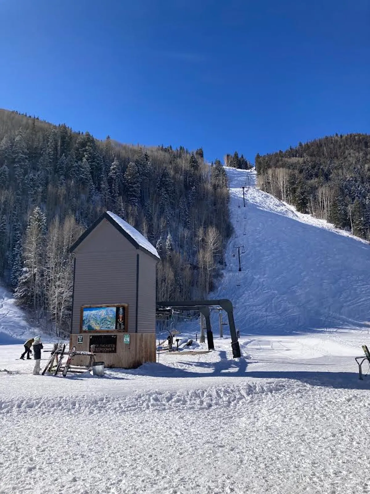 A classic winter scene with a ski lift ascending over snow-covered terrain, a ticket booth, and winter trees framing the mountain slope. The crisp snow and active ski environment evoke the thrill and convenience of immediate access to the slopes.