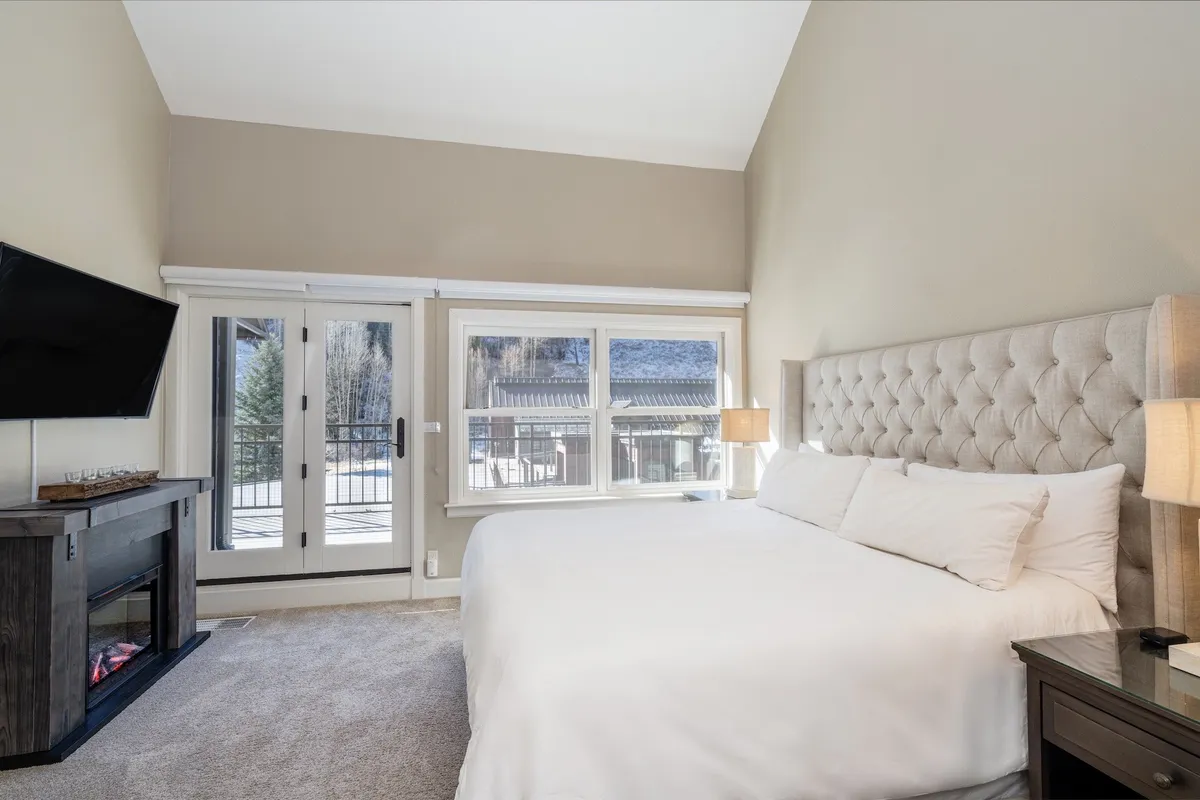 A beautifully appointed primary bedroom with a king-sized bed, large windows that fill the space with natural light, an electric fireplace for added warmth, and a wall-mounted television. The ambiance is cozy and serene, suggesting a perfect spot for romance or restful sleep.
