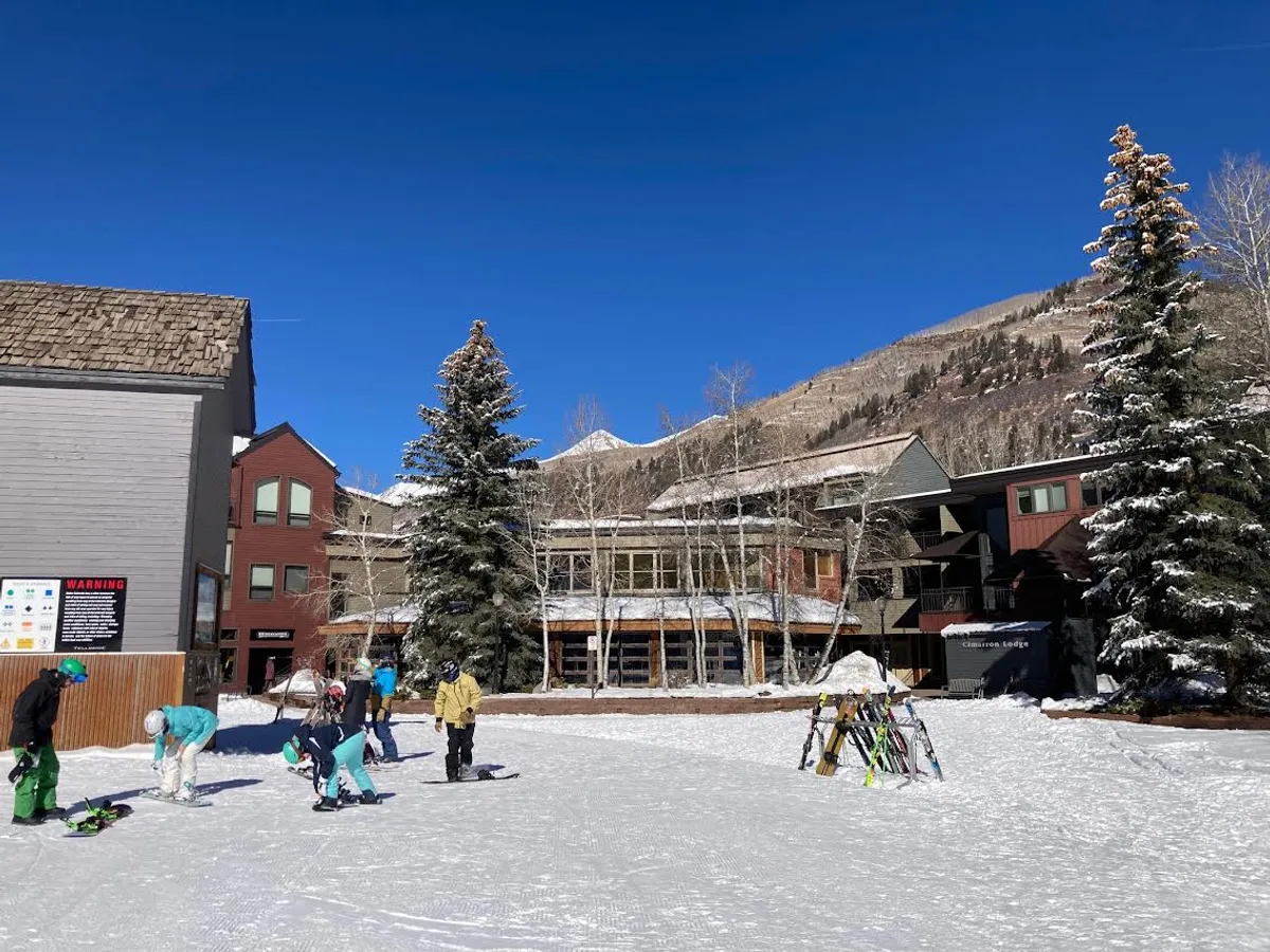 People prepare for a day of skiing in a snowy, slope-side locker and staging area, with nearby trees and lodge buildings in view. The atmosphere buzzes with anticipation and camaraderie, capturing the convenience of direct ski access.