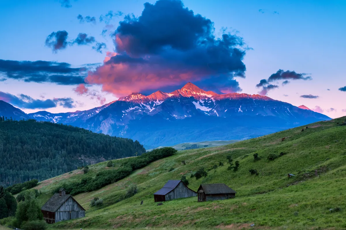 A sweeping view of Telluride’s iconic mountains, rustic barns, and lush green hills beneath a dramatic sky. The tranquil landscape invites quiet moments of reflection and a deep sense of place.
