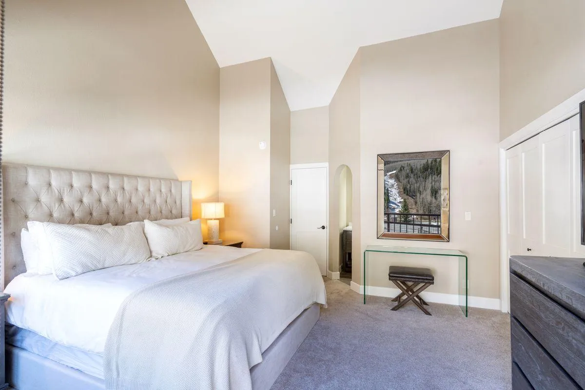 A serene bedroom with a king-sized bed, decorative mirror, and transparent console table set against a backdrop of natural light from large windows. The view outside hints at access to an outdoor space, aligning with the suite's deck access feature.