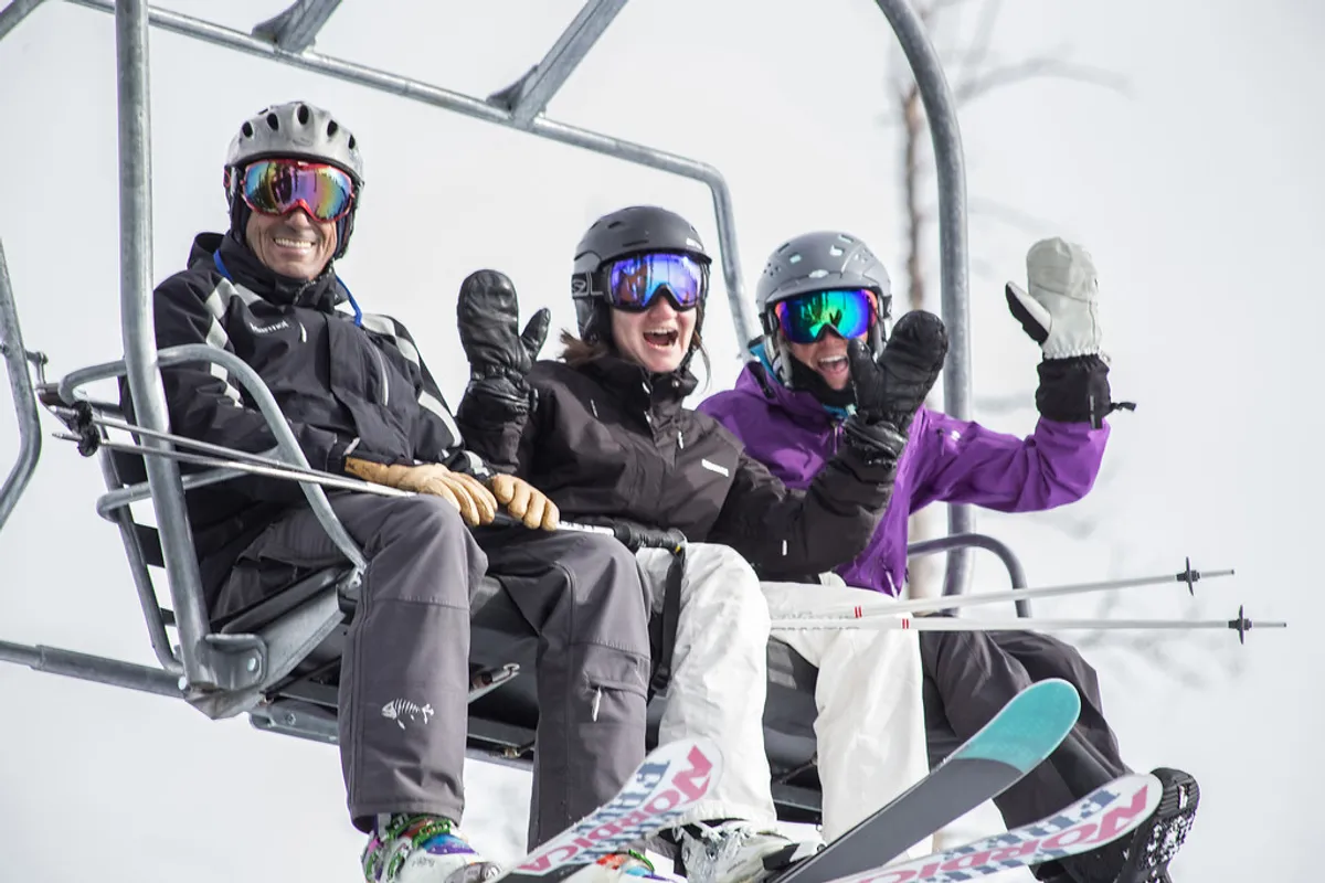 This image shows three skiers riding a ski lift over a snowy landscape, with ski equipment in tow and distant mountain vistas. The cheerful mood and sense of camaraderie highlight the thrill and social aspect of a day on the slopes.