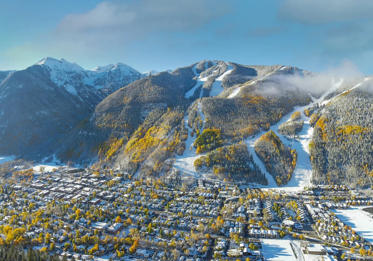 Sweeping view of Telluride featuring snow-capped mountains, ski slopes, the town nestled below, and vivid autumn foliage. The scene captures both the thrill of winter activities and the vibrant beauty of the fall season.