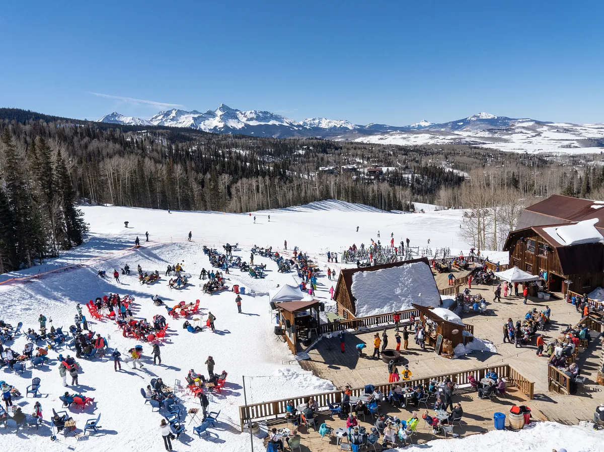 A vibrant scene at the ski slopes: skiers and snowboarders glide down sunlit hills, a crowd enjoys après-ski gatherings outside a lodge with mountain ranges in the background. The lively mood and outdoor seating underscore relaxation and community following a day of adventure.