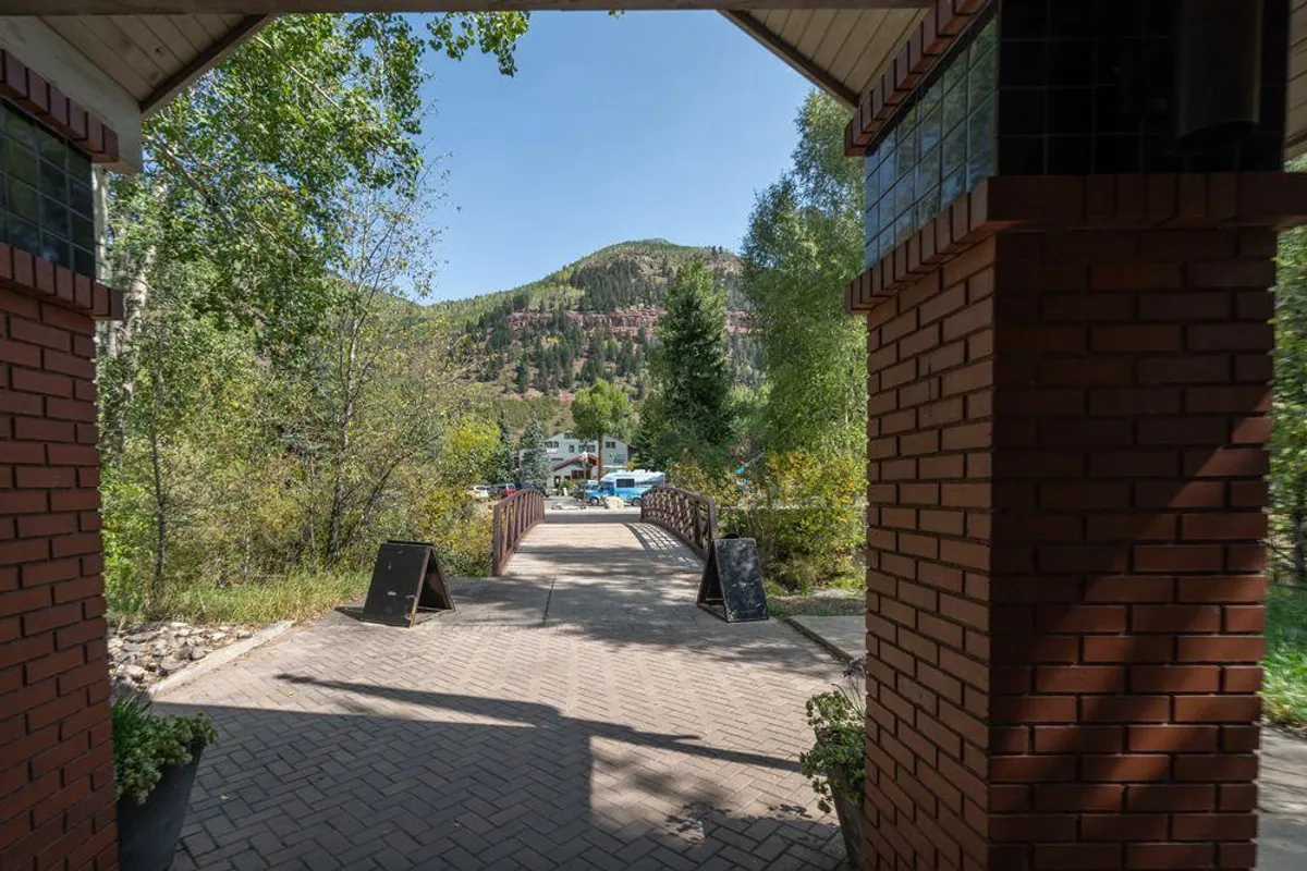 A serene exterior scene featuring an arched stone entranceway leading onto a charming bridge, surrounded by lush greenery. The pathway invites leisurely nature walks and exploration, while the tranquil environment evokes relaxation amidst Telluride’s natural beauty.