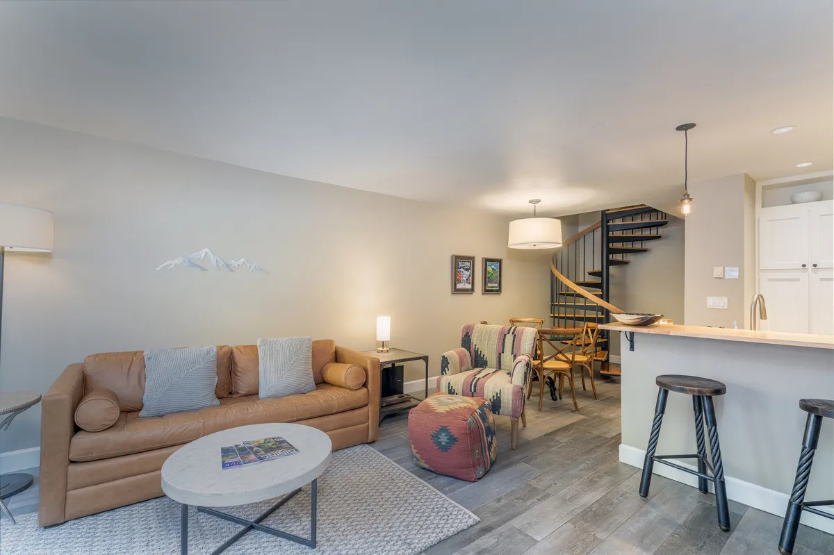 A welcoming living room featuring a leather sofa, colorful armchairs, a spiral staircase, and a bar counter. Ample natural light streams through large picture windows, creating a bright and inviting atmosphere. The arrangement encourages relaxation and conversation, and the open-plan design provides a seamless flow into adjacent spaces.