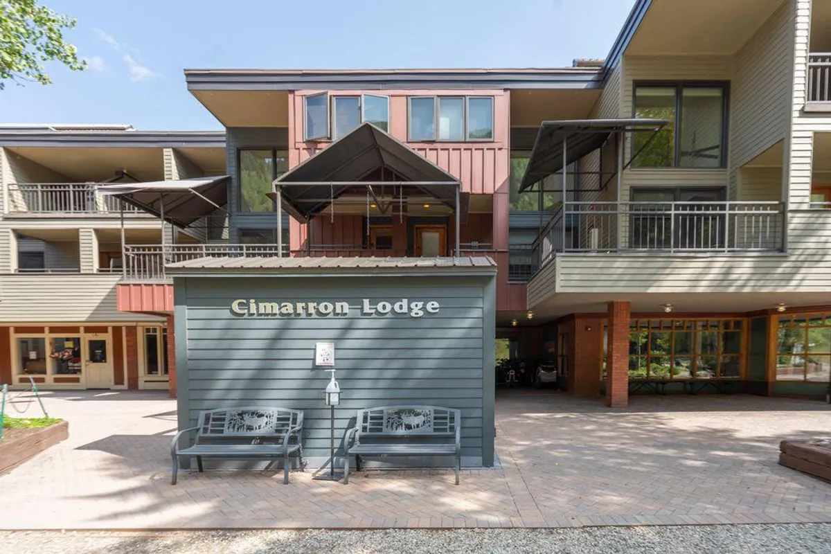 The welcoming exterior of Cimarron Lodge features an inviting entrance, benches for relaxing outdoors, and subtle touches of local character. The lodge is framed by landscaping and mountain views beyond.